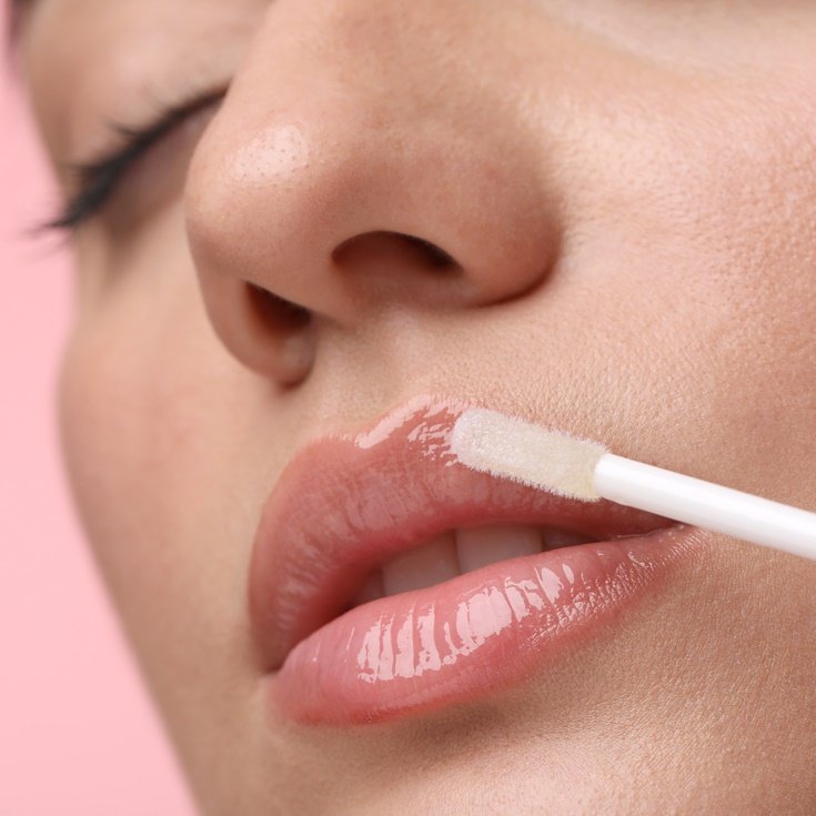 Mulher aplicando brilho labial em fundo rosa.