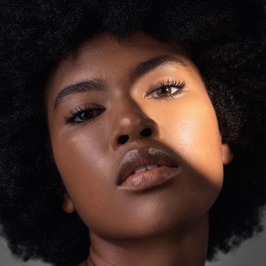 Um close-up mostra o rosto de uma mulher negra com cabelo afro volumoso. A luz incide sobre sua pele, destacando os detalhes de sua maquiagem.