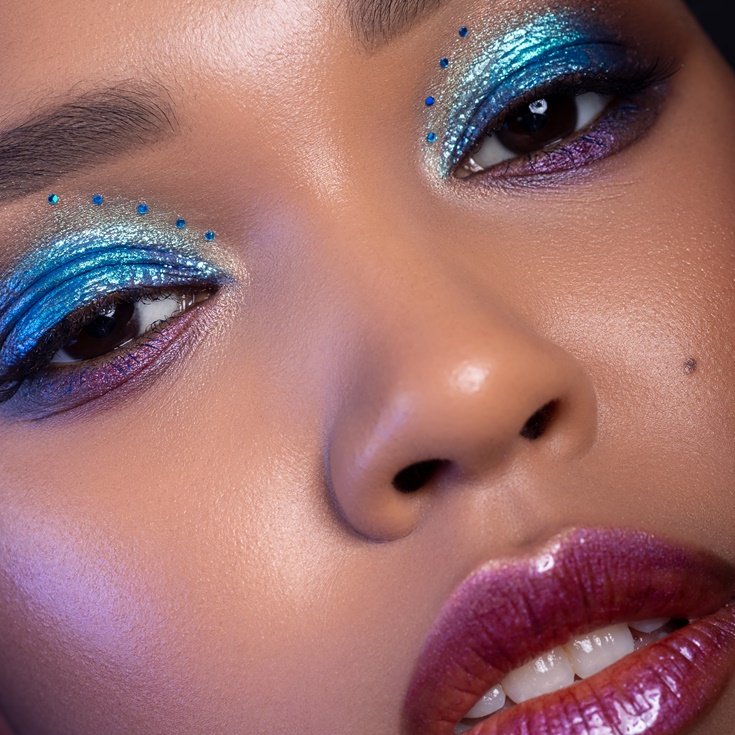 Maquiagem artística e colorida para festa de ano novo com sombra azul e roxa brilhante, pedrarias delicadas e batom roxo metálico, destacando as cores do ano novo de forma ousada