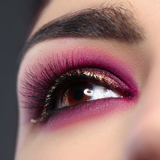 Close-up de um olho com maquiagem rosa e brilho, mostrando a aplicação detalhada da sombra e dos cílios postiços.