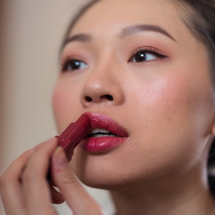 Mulher asiática passando batom rosa para completar sua maquiagem soft glam.