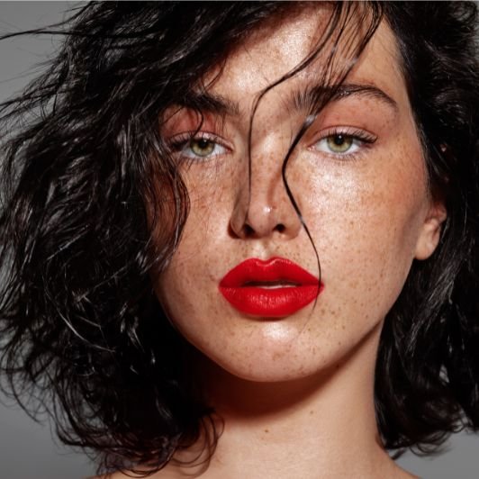 Mulher com batom vermelho tomate nos lábios, olhos verdes e cabelo preto curto com ondas.