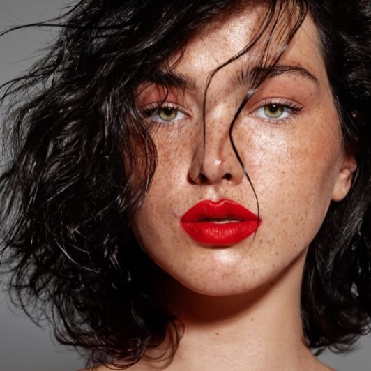 Mulher com batom vermelho tomate nos lábios, olhos verdes e cabelo preto curto com ondas.
