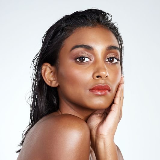 Mulher com cabelo molhado e mão no rosto enquanto usa batom coral nos lábios.