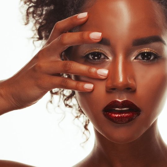 Mulher com a mão no rosto e maquiagem dourada nos olhos e batom vermelho cintilante.