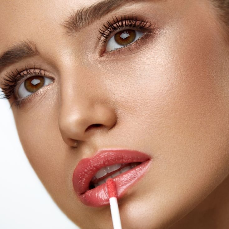 Close nos lábios femininos com gloss labial rosa aplicado com pincel de maquiagem. Os lábios estão intensamente brilhantes, com efeito espelhado e acabamento volumoso, evidenciando hidratação e cor uniforme