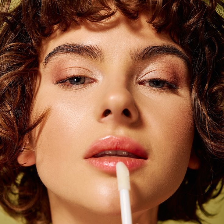 Mulher passando lip oil nos lábios em fundo verde.