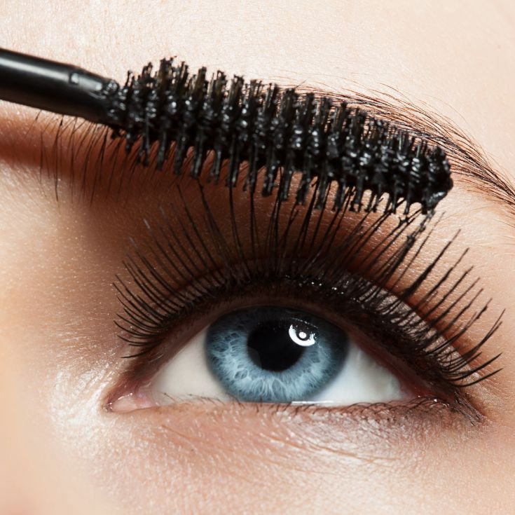 Mulher com olhos azuis aplicando rímel nos seus cílios.