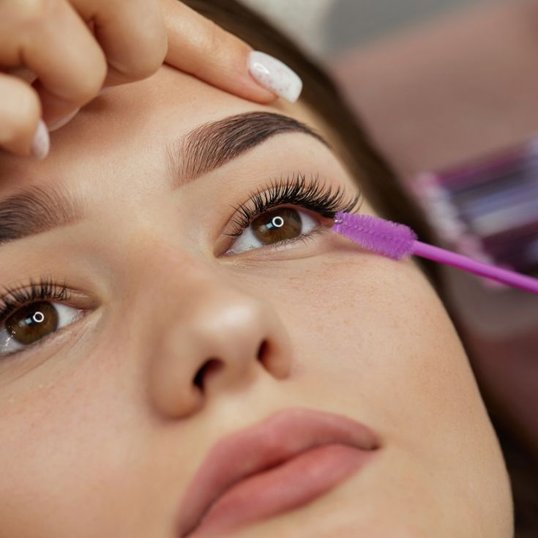 Cliente olhando para frente enquanto a esteticista penteia seus cílios fio a fio com uma escovinha roxa