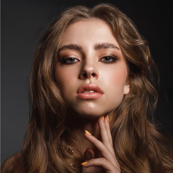 Mulher com maquiagem em tom marrom no côncavo, batom nude nos lábios enquanto toca o queixo com os dedos.