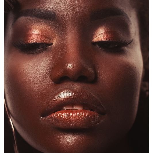 Mulher negra com maquiagem marrom em tons metálicos na pálpebra e lábios com gloss. 