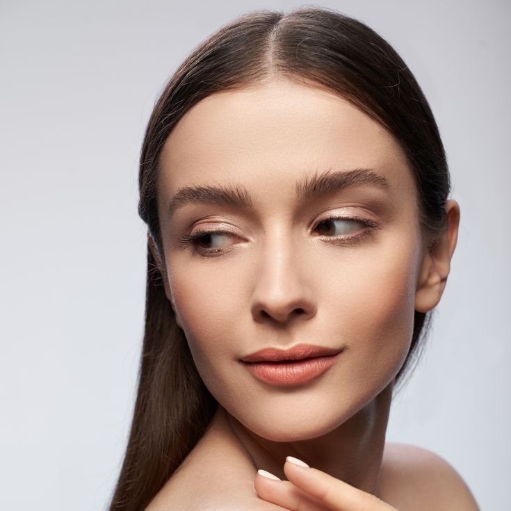 Mulher com a mão no ombro olhando para à esquerda com maquiagem leve no rosto com contorno facial e base matte.