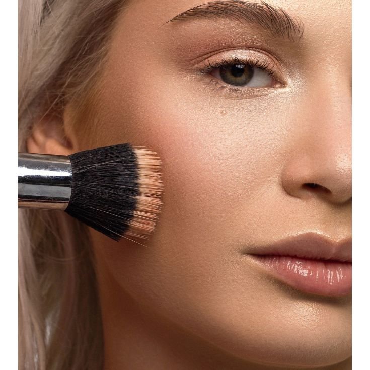 Menina olhando para a câmera enquanto aplica base matte na sua bochecha com pincel duo fiber.