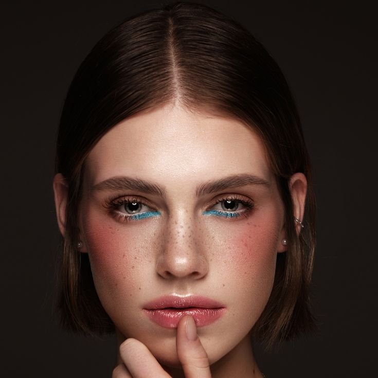 Menina com o dedão na boca enquanto usa sombra azul na parte inferior dos olhos e blush em tom pêssego.