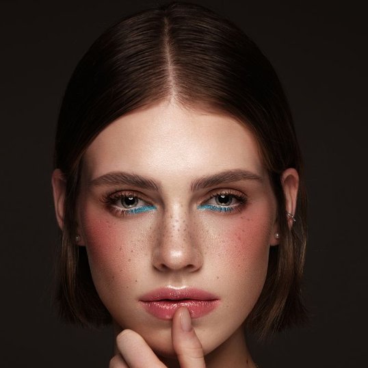 Menina com o dedão na boca enquanto usa sombra azul na parte inferior dos olhos e blush em tom pêssego.