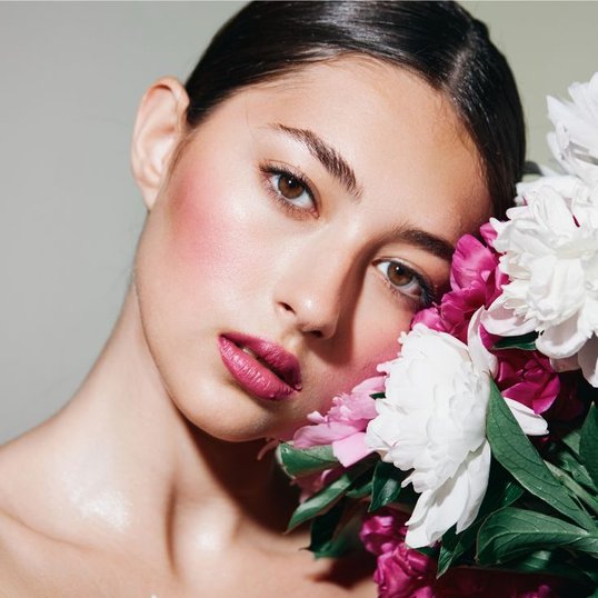 Mulher encostando sua cabeça em buquê de flores branco e rosa. Ela usa blush em tom rosa terroso e batom rosa nos lábios.