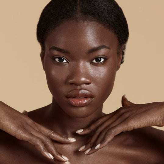 Mulher com rosto oval e mãos sobrepostas no seu ombro. Sua pele está com aspecto natural e com pontos iluminados.