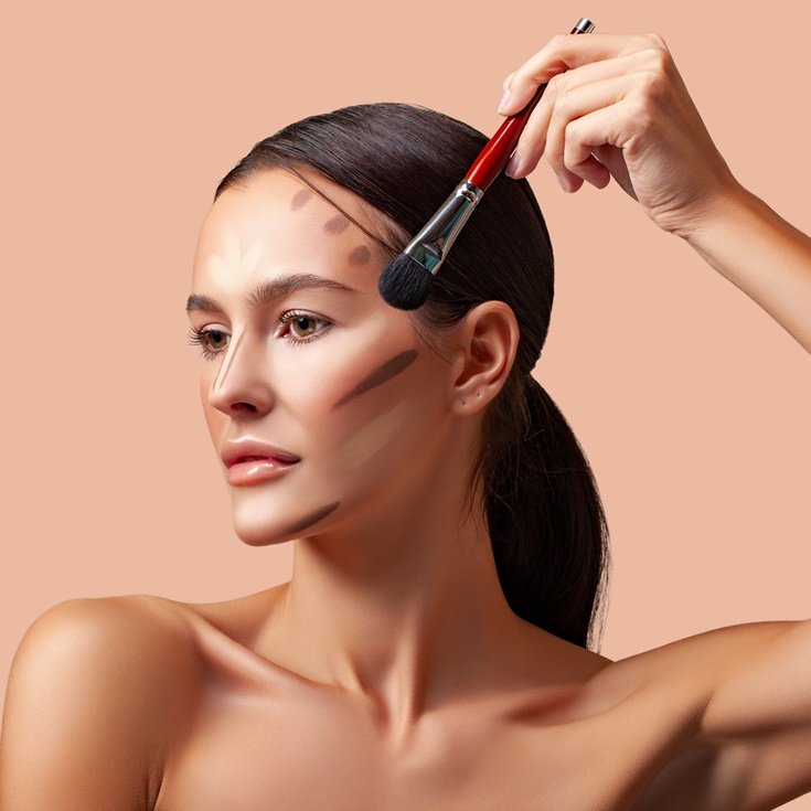 Mulher aplicando contorno no rosto com pincel em fundo bege.