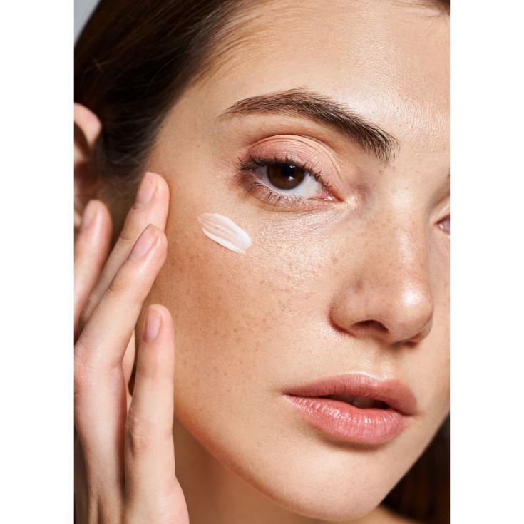 Close-up de uma pessoa aplicando hidratante facial na bochecha. A imagem mostra parte do rosto com pele natural, uma pequena quantidade de produto branco sendo espalhado e dedos tocando delicadamente a face.