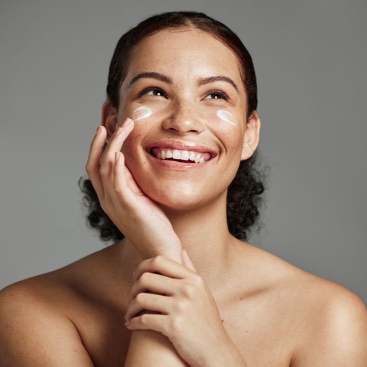 Mulher sorridente aplicando hidratante facial na bochecha com o dedo. A pele apresenta aparência luminosa com pontos de produto branco sendo espalhados.