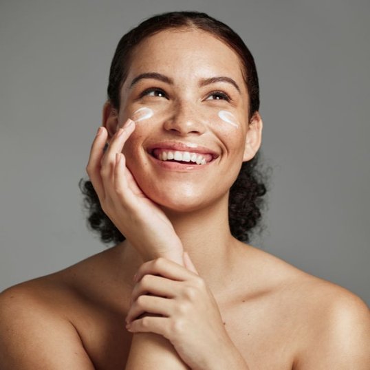 Mulher sorridente aplicando hidratante facial na bochecha com o dedo. A pele apresenta aparência luminosa com pontos de produto branco sendo espalhados.