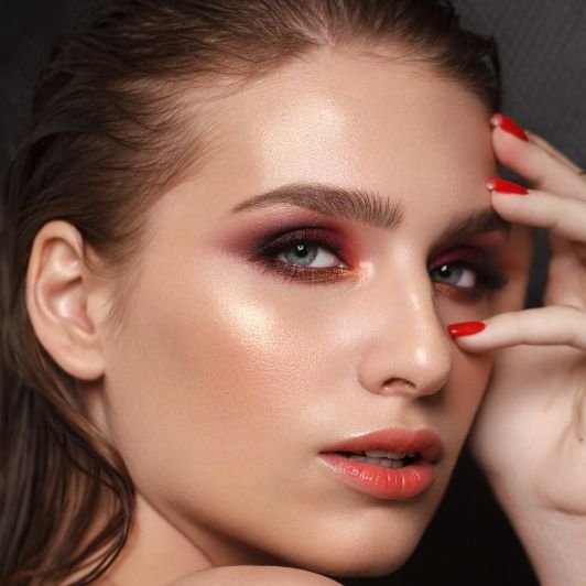 Mulher com dedos no rosto usando maquiagem colorida nos olhos e iluminador champagne nas bochechas