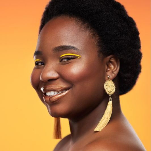 Mulher sorridente com delineador gráfico amarelo nos olhos, brincos longos amarelos e iluminador líquido dourado nas bochechas.
