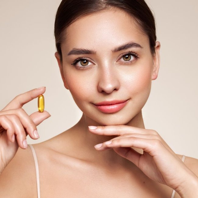 Mulher com uma mão no queixo e a outra mão segurando uma cápsula de vitamina E.