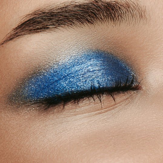 Close-up da pálpebra com a sombra colorida em tons de azul vibrante. A sombra se estende do canto interno ao externo do olho, criando um visual marcante.