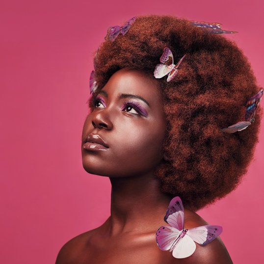 Mulher negra com um afro volumoso decorado com borboletas roxas, exibindo uma maquiagem vibrante para o Carnaval.