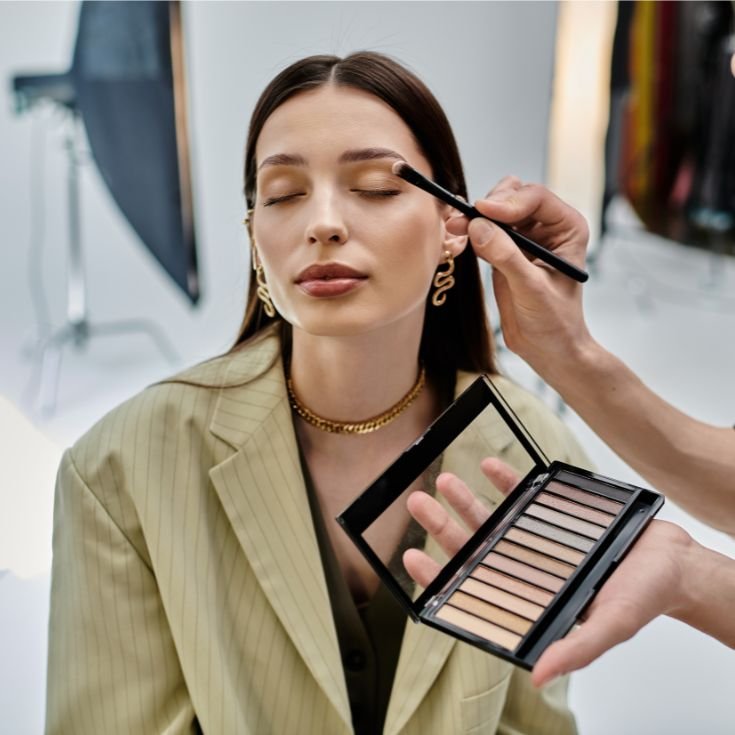 Maquiadora aplicando sombra marrom no côncavo de sua cliente, que está com os olhos fechados.