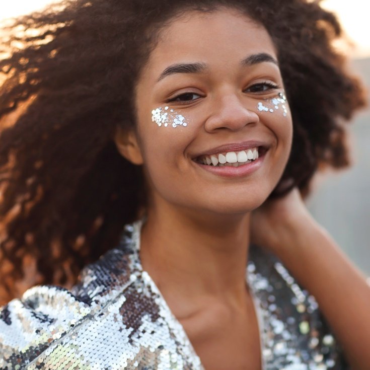 Mulher sorridente com maquiagem de Carnaval, com glitter prata aplicado no rosto e roupa brilhante, aproveitando a folia.