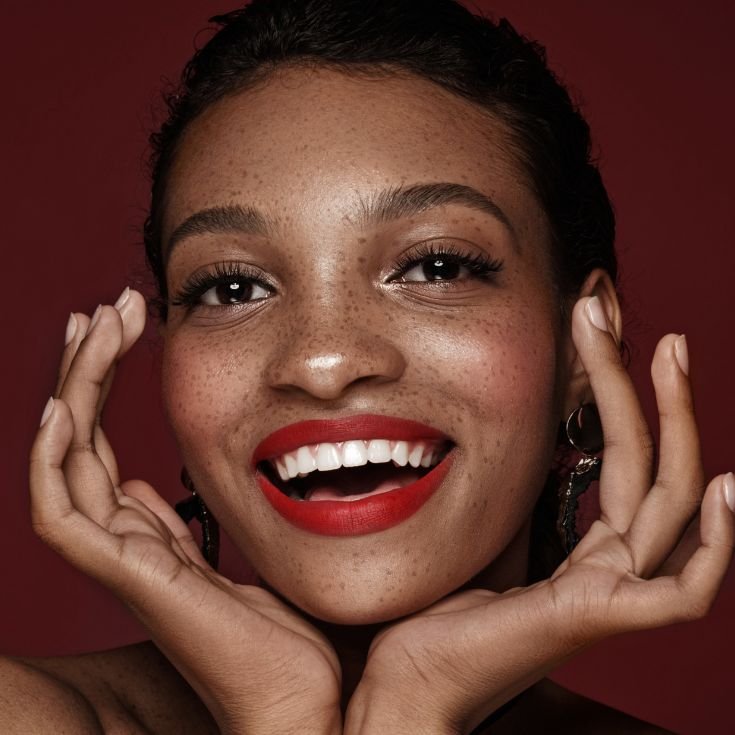 Mulher com as mãos no rosto e maquiagem simples nos olhos com batom vermelho.