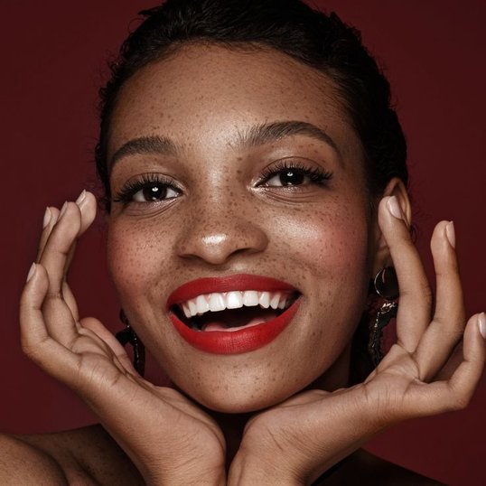 Mulher com as mãos no rosto e maquiagem simples nos olhos com batom vermelho.