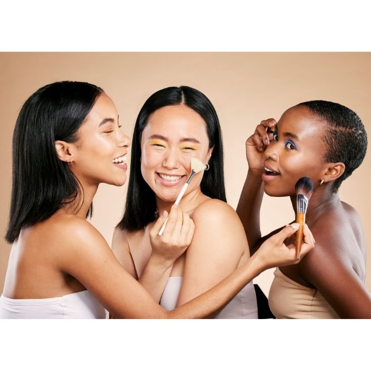 Três mulheres de etnias diferentes se aplicam maquiagem e riem.