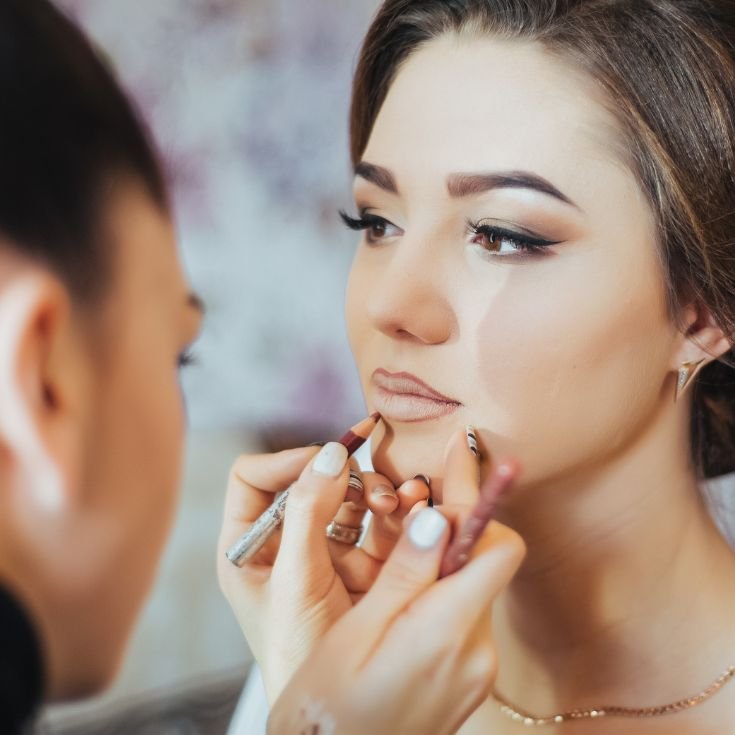 Noiva sendo maquiada por maquiadora que está passando lápis labial em sua boca.