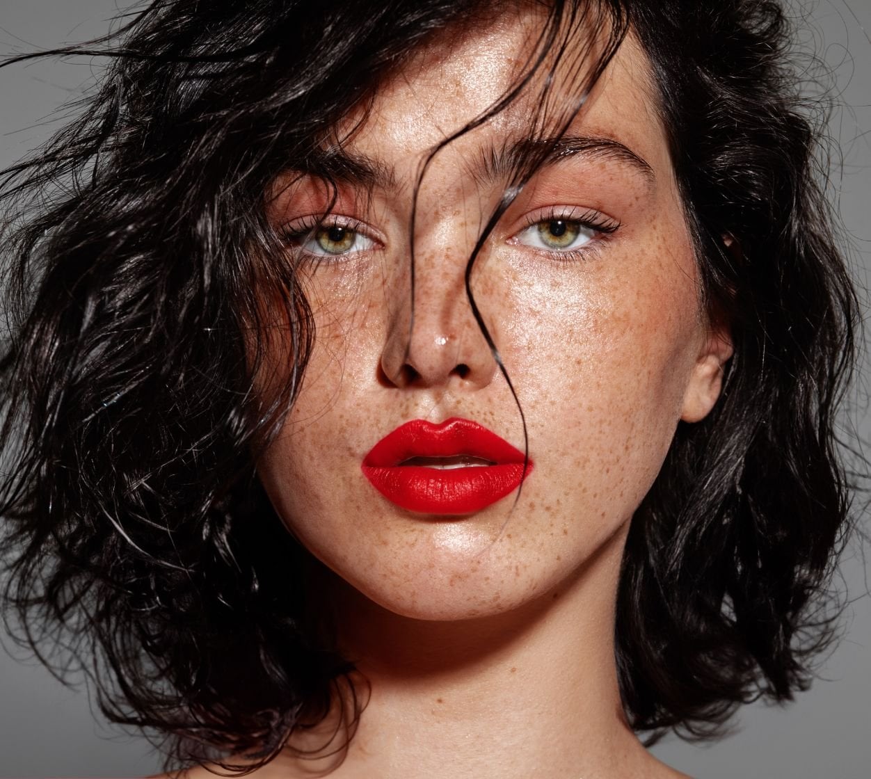 Mulher com batom vermelho tomate nos lábios, olhos verdes e cabelo preto curto com ondas.
