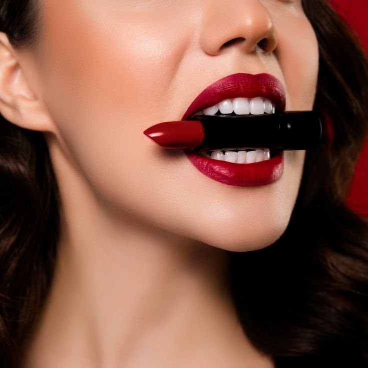 Mulher usando batom vermelho escuro nos lábios enquanto segura um batom em sua boca