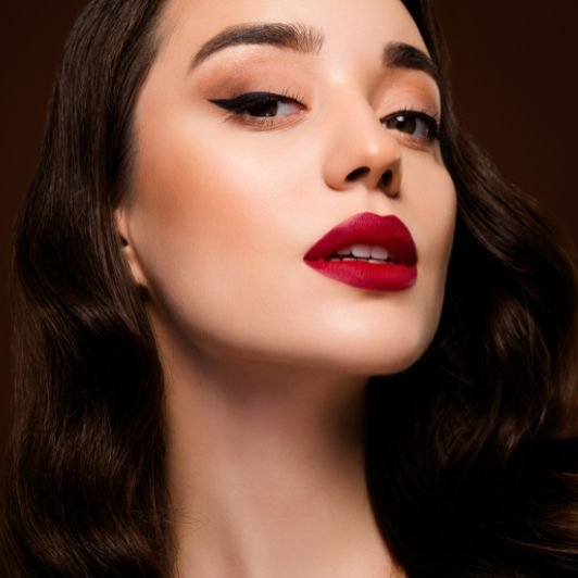 Mulher branca usando um tom de batom vermelho escuro, vestido preto e maquiagem dos olhos simples.