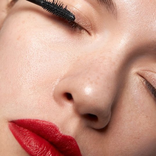 Mulher de olhos fechados usa máscara de cílios para aplicar maquiagem nos olhos que combine com seu batom vermelho.