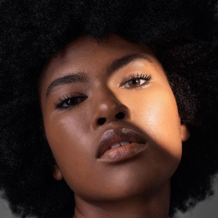 Mulher com pele naturalmente iluminada, com gloss translúcido e rímel preto com o rosto iluminado parcialmente pelo sol.