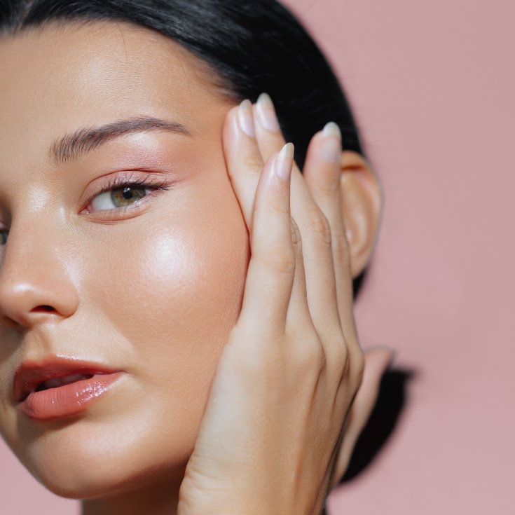 Mulher de pele clara com maquiagem fresh glow e mão no rosto em fundo rosa.