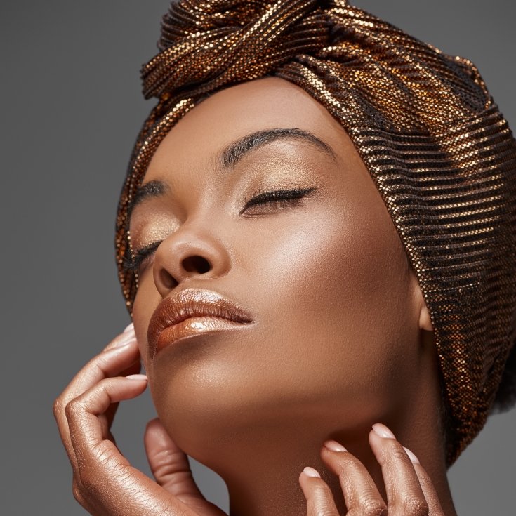 Mulher com pele radiante, olhos fechados e turbante dourado em fundo cinza.