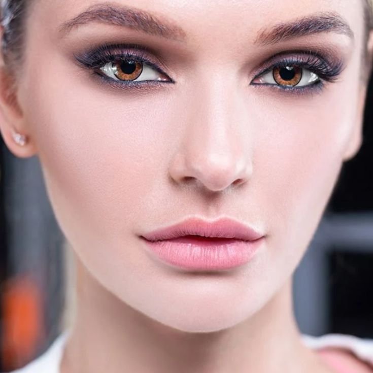 Mulher com maquiagem estilo fox eyes, sombra esfumada em tons escuros azulados, lábios rosados naturais e brinco discreto.