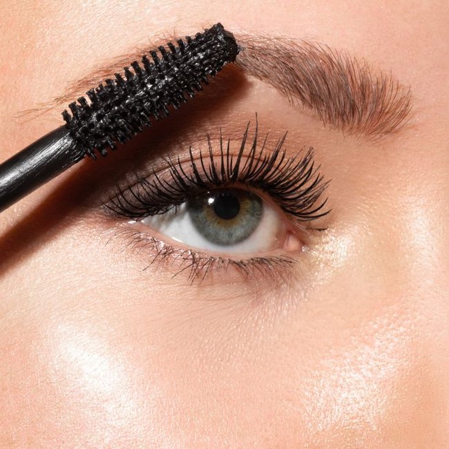 Close em olhos verdes com cílios volumosos e definidos sendo maquiados com rímel preto e sobrancelhas naturais em destaque.