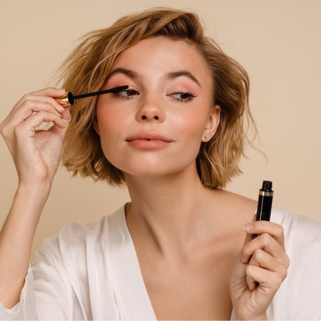 Mulher loira de cabelo curto aplica rímel no olho esquerdo, segurando o tubinho preto. Usa roupão branco, maquiagem rosada, fundo bege.