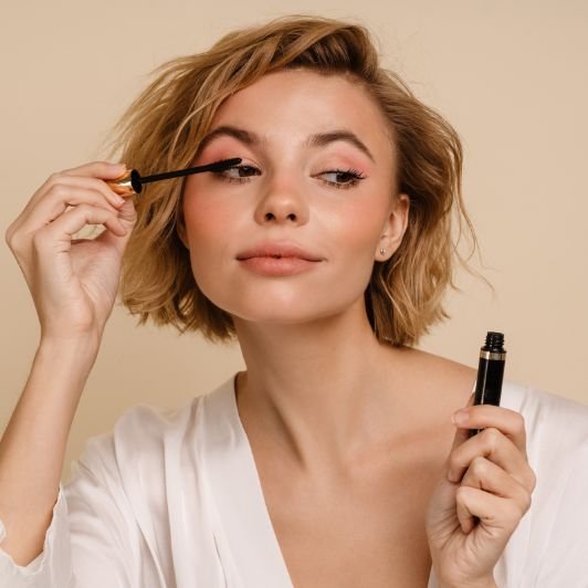Mulher jovem de cabelo curto loiro aplicando máscara de cílios com um olhar concentrado e maquiagem suave em tons rosados sobre fundo bege neutro.
