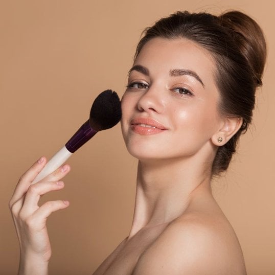 Jovem mulher sorridente aplica o blush em pó com um pincel aplicador