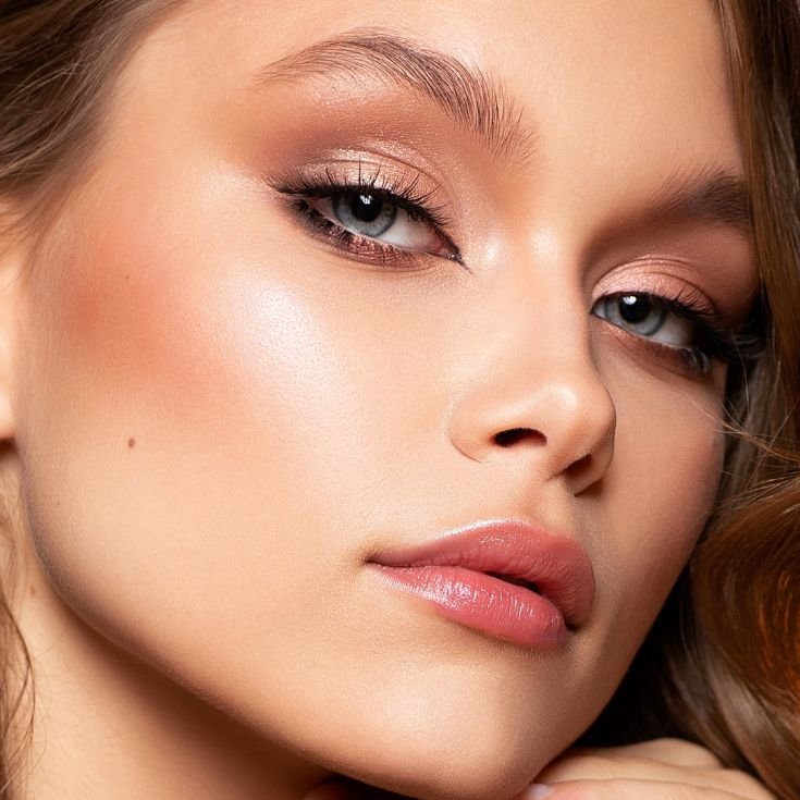 Mulher jovem com olhos azuis, maquiagem smoky rose gold, delineado gatinho, blush suave, lábios rosados glossy e pele iluminada.