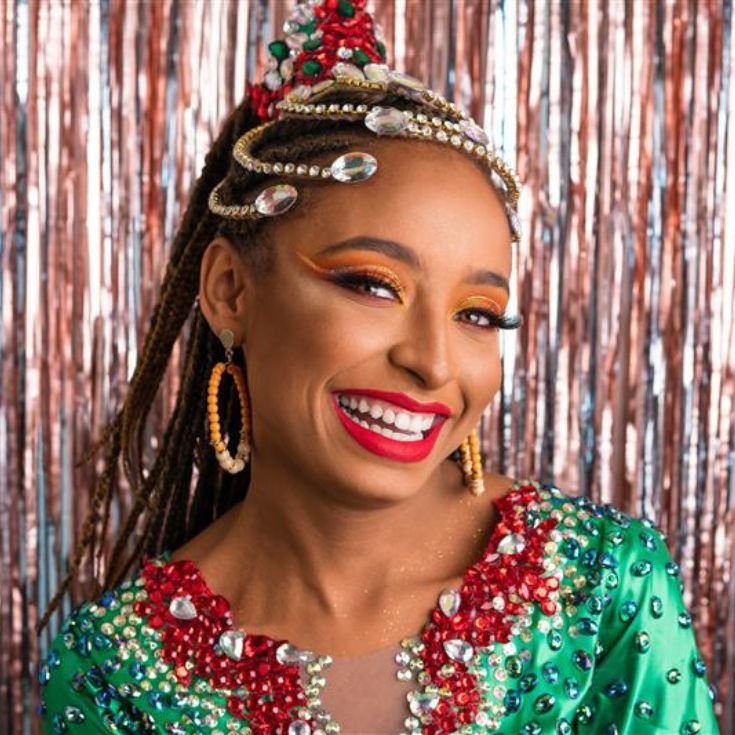 Uma mulher sorridente com uma elaborada maquiagem de carnaval, incluindo sombras de olhos vibrantes em tons de laranja e amarelo, delineador marcante e cílios postiços.